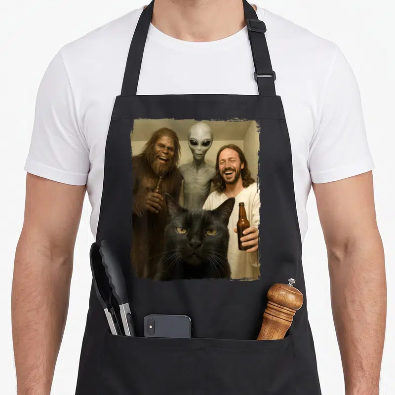 Jesus funny cat apron - Black cat foreground with laughing Bigfoot, grey alien, and Jesus holding brown bottles — shown on a black cotton canvas apron against a white background