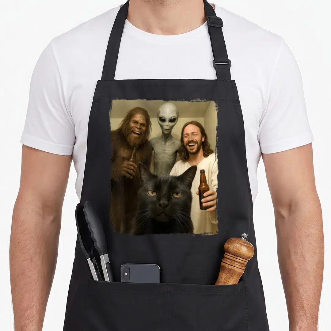 Jesus funny cat apron - Black cat foreground with laughing Bigfoot, grey alien, and Jesus holding brown bottles — shown on a black cotton canvas apron against a white background