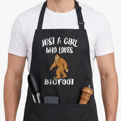 Sasquatch bigfoot apron for women - Brown cartoon bigfoot centered between white text reading Just A Girl Who Loves — shown on a black cotton canvas apron against a white background