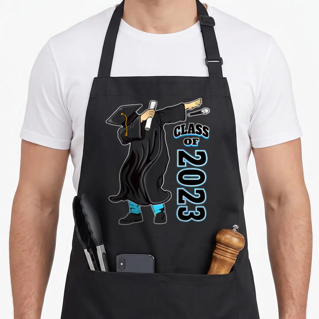 School senior apron - Cartoon graduate dabbing in black gown holding diploma next to vertical Class of 2023 text — shown on a black cotton canvas apron against a white background