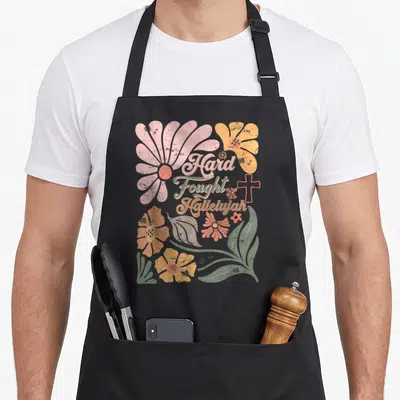 Christian boho apron - Retro pink and yellow flowers surrounding stacked Hard Fought Hallelujah text with a small cross — shown on a black cotton canvas apron against a white background