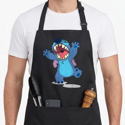 Growl stitch apron - Blue cartoon creature with open mouth, purple ears, and white puddle beneath feet — shown on a black cotton canvas apron against a white background