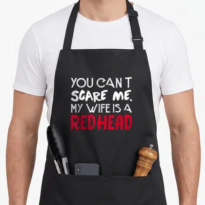 Redhead wife apron - Stacked white and red text reading You Can't Scare Me My Wife Is A Redhead — shown on a black cotton canvas apron against a white background