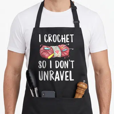 Unravel crocheter crochet apron - I CROCHET SO I DON'T UNRAVEL text over pink yarn ball with hooks and heart label — shown on a black cotton canvas apron against a white background
