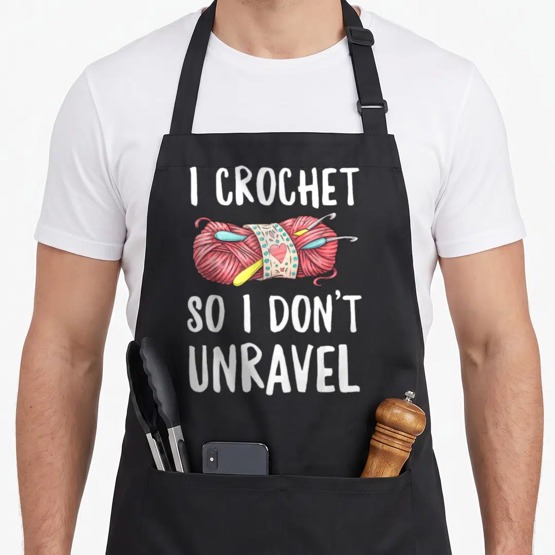 Unravel crocheter crochet apron - I CROCHET SO I DON'T UNRAVEL text over pink yarn ball with hooks and heart label — shown on a black cotton canvas apron against a white background