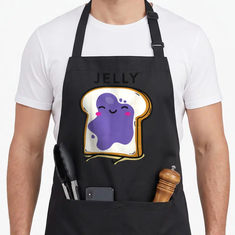 Couple butter apron - Cartoon toast with purple jelly blob and JELLY text in black lettering — shown on a black cotton canvas apron against a white background