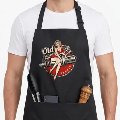 Rod car apron - Old School REPAIR text with woman in red dress sitting on shock absorber inside chain-bordered circle — shown on a black cotton canvas apron against a white background