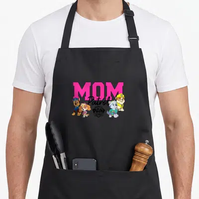 Cute patrol happy apron - MOM Patrol text with four cartoon puppies in pink blue yellow and black — shown on a black cotton canvas apron against a white background