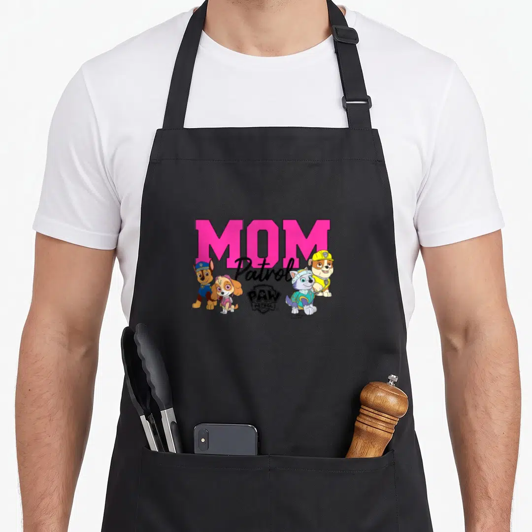 Cute patrol happy apron - MOM Patrol text with four cartoon puppies in pink blue yellow and black — shown on a black cotton canvas apron against a white background