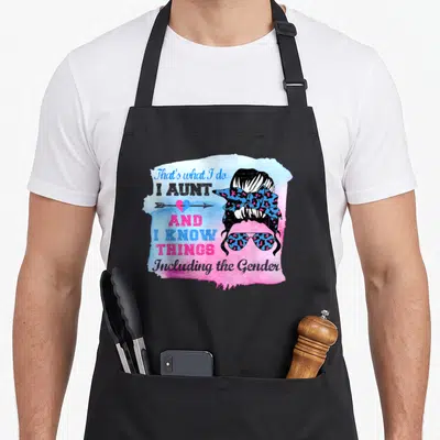Keeper i love buns apron - Cartoon woman head with sunglasses and headband next to text about knowing gender on blue — shown on a black cotton canvas apron against a white background