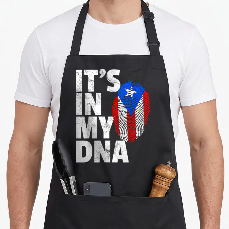 Dna rican apron - Distressed white IT'S IN MY DNA text next to a red white and blue fingerprint shaped like the Puerto — shown on a black cotton canvas apron against a white background