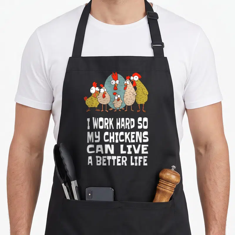 Hard chicken apron - Five cartoon chickens and a large blue egg below white text reading I work hard so my chickens can — shown on a black cotton canvas apron against a white background