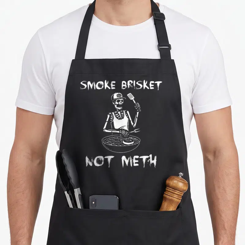 Smoke skeleton apron - White skeleton wearing BBQ hat holding spatula and fork behind grill with text Smoke Brisket Not — shown on a black cotton canvas apron against a white background