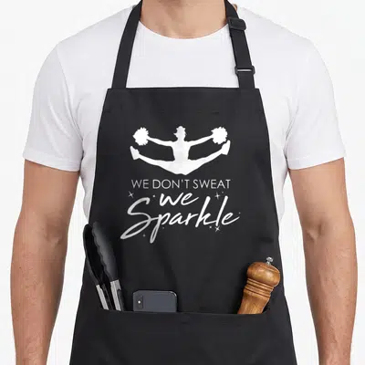 Cheerleading cheers apron - White jumping cheerleader silhouette above We Don't Sweat we Sparkle text with small — shown on a black cotton canvas apron against a white background