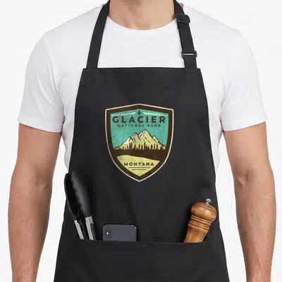 National glaciers apron - Retro shield badge with mountains and Glacier National Park Montana text in brown teal beige — shown on a black cotton canvas apron against a white background