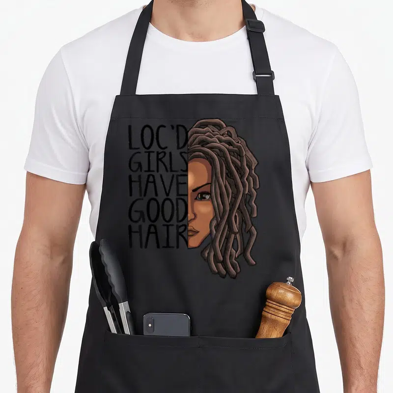 Loc hair apron - Loc'd Girls Have Good Hair text beside cartoon woman face with brown dreadlocks — shown on a black cotton canvas apron against a white background