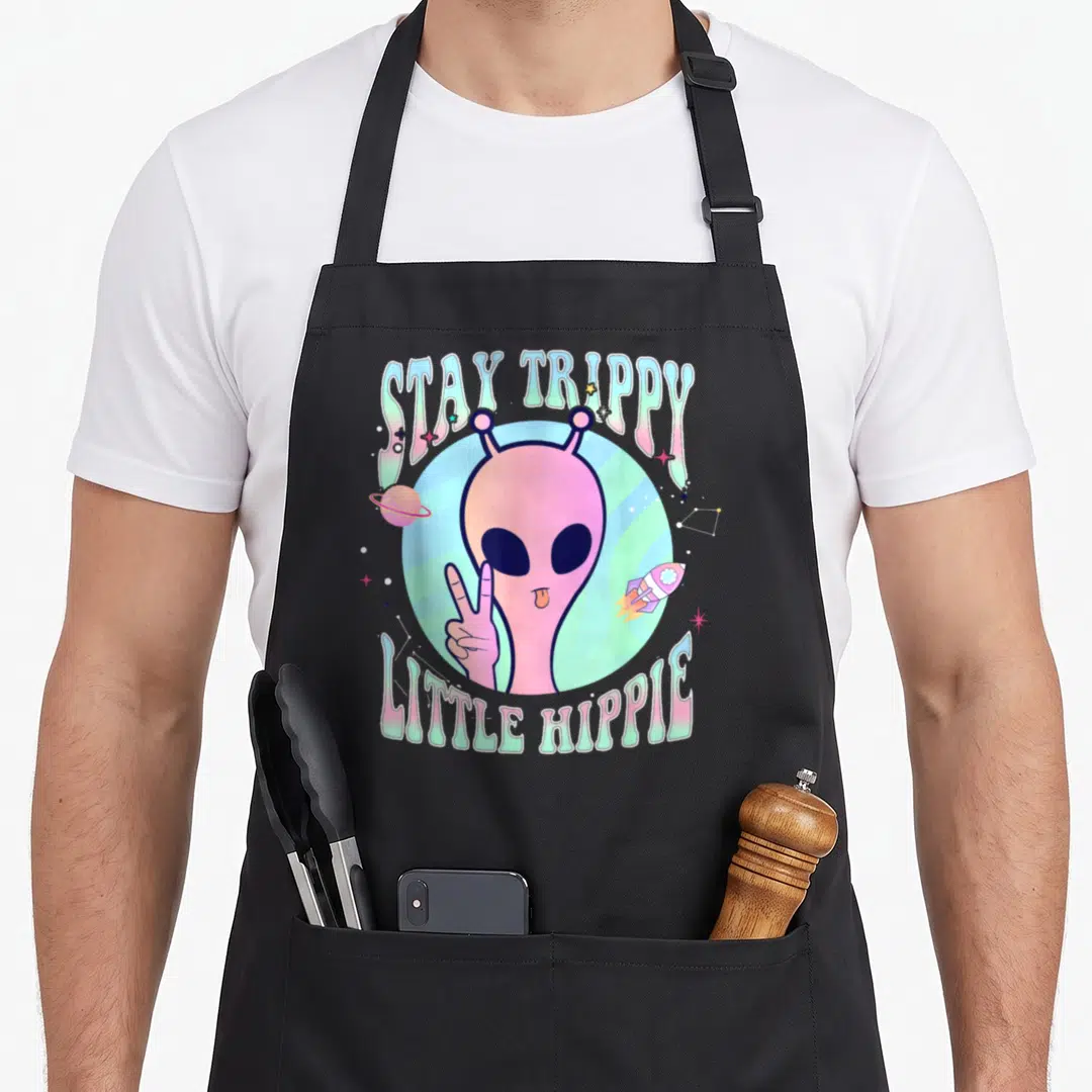 Trippy hippie apron - Pink alien holding peace sign with STAY TRIPPY LITTLE HIPPIE text and space objects — shown on a black cotton canvas apron against a white background