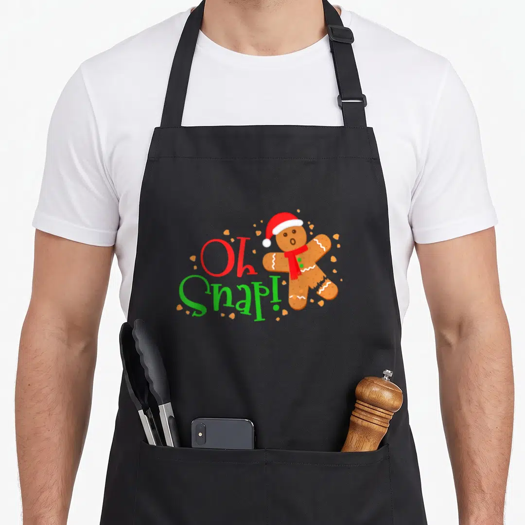 Snap holiday baking apron - Broken gingerbread man with Santa hat next to Oh Snap text and scattered crumbs — shown on a black cotton canvas apron against a white background