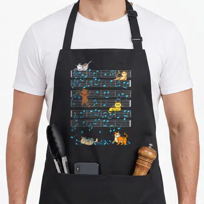 Playing kitty cat apron - Five cartoon cats in blue orange and brown on musical staff lines — shown on a black cotton canvas apron against a white background