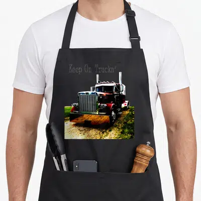 Youth semi truck apron - Keep On Truckn' text above red semi-truck photo on dirt and grass — shown on a black cotton canvas apron against a white background