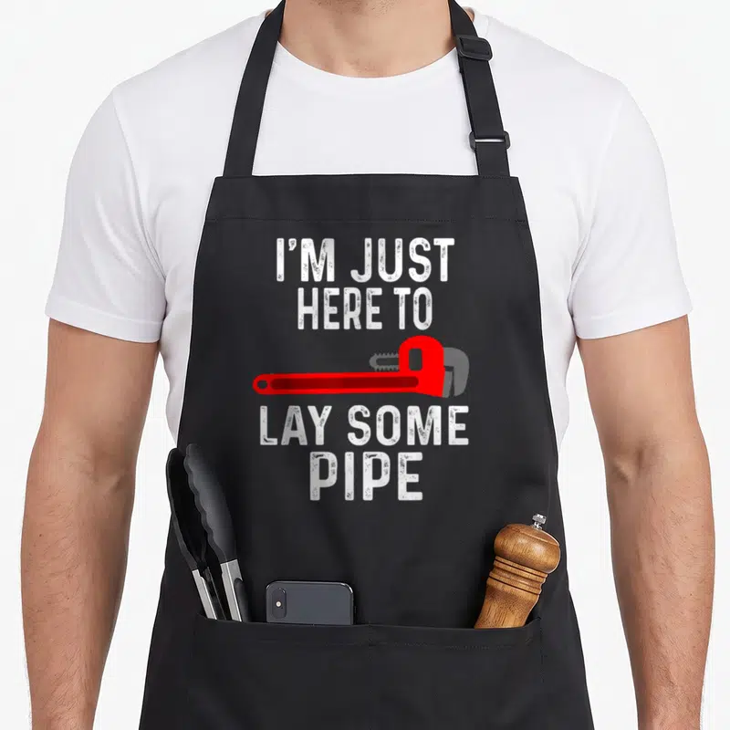 Plumbing pipe apron - Red pipe wrench between white text reading I'M JUST HERE TO LAY SOME PIPE — shown on a black cotton canvas apron against a white background