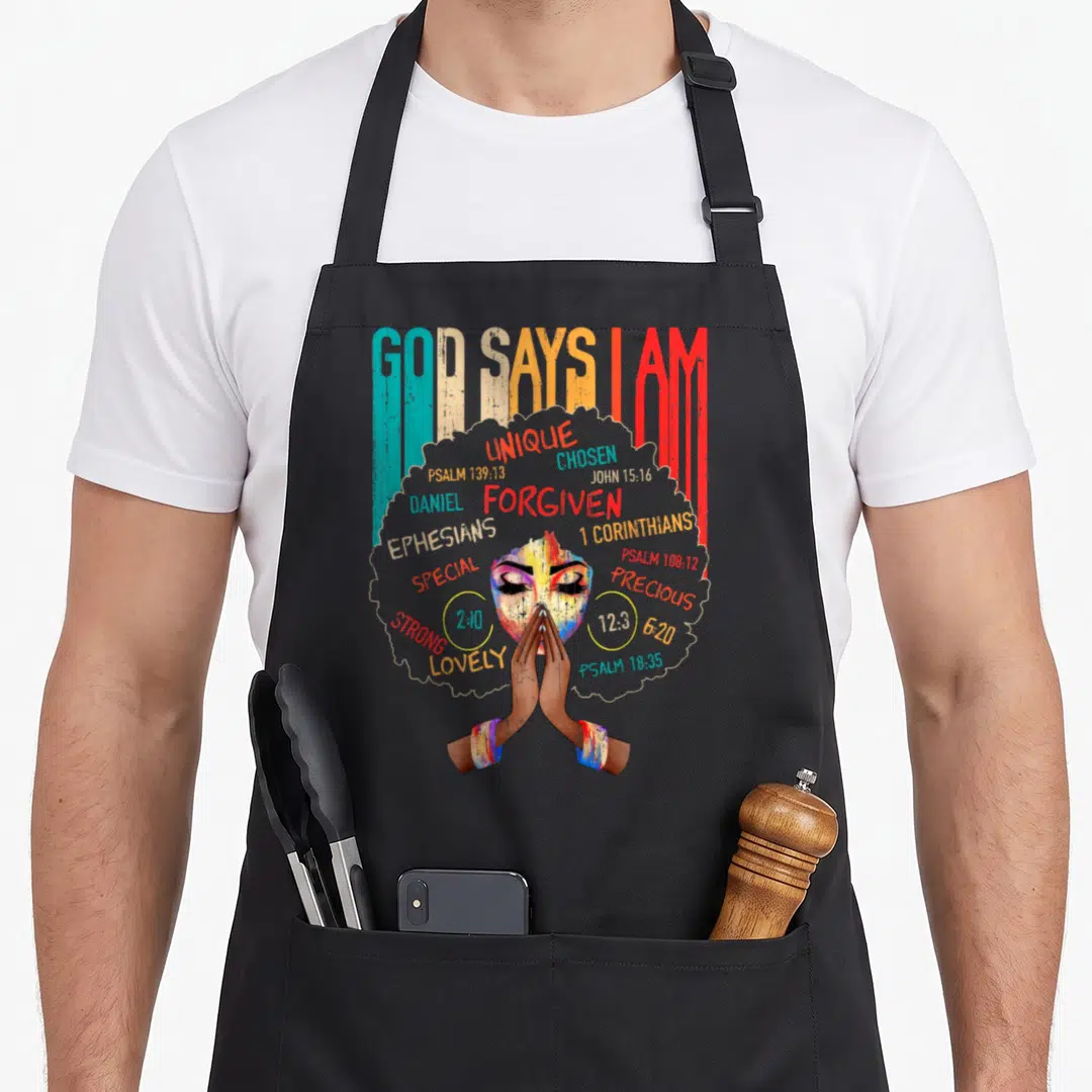Melanin god says i am apron - GOD SAYS I AM text above praying woman face surrounded by biblical words in watercolor — shown on a black cotton canvas apron against a white background