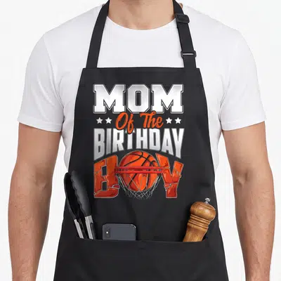 Basketball family aprons - MOM Of The BIRTHDAY BOY text with basketball going through red hoop and orange stars — shown on a black cotton canvas apron against a white background