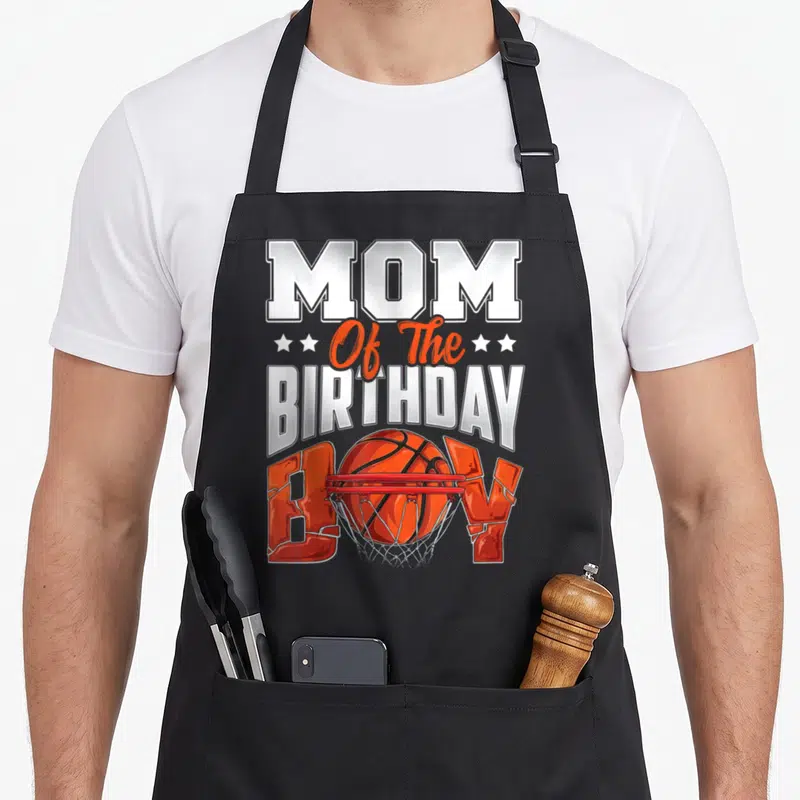 Basketball family aprons - MOM Of The BIRTHDAY BOY text with basketball going through red hoop and orange stars — shown on a black cotton canvas apron against a white background