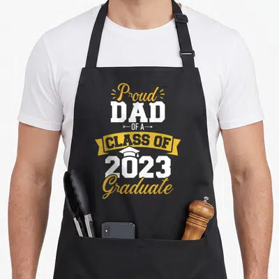 Graduate class dad apron - Stacked yellow white and blue text reading proud dad of a class of 2023 graduate with small — shown on a black cotton canvas apron against a white background