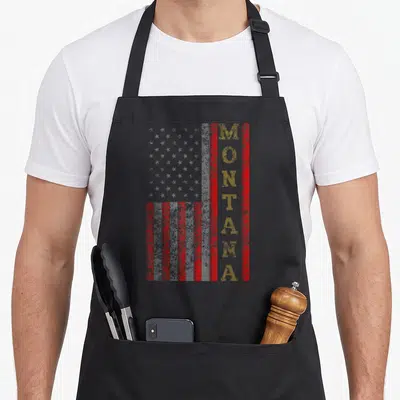 Montana patriotic apron - Vertical MONTANA text in yellow overlapping a distressed red and white dotted US flag pattern — shown on a black cotton canvas apron against a white background
