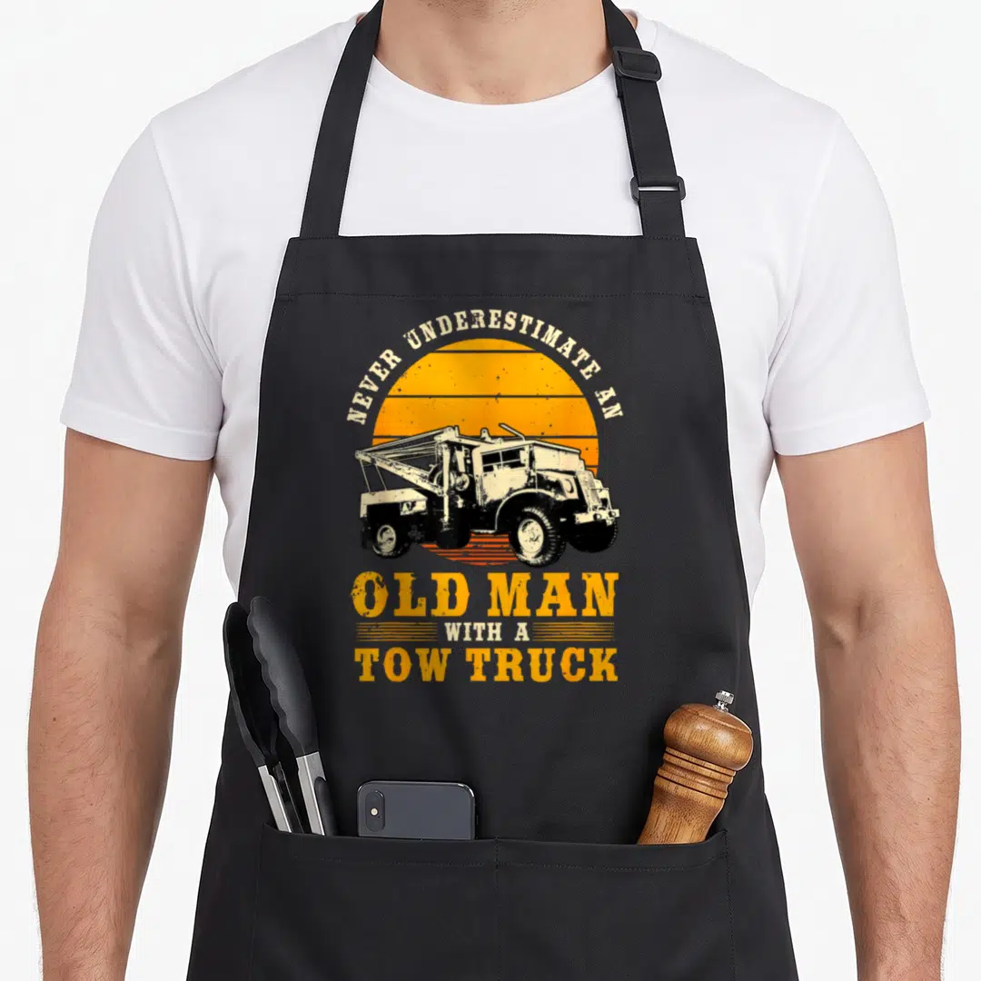 Underestimate truck apron - Retro tow truck illustration with orange striped sun and arched text — shown on a black cotton canvas apron against a white background