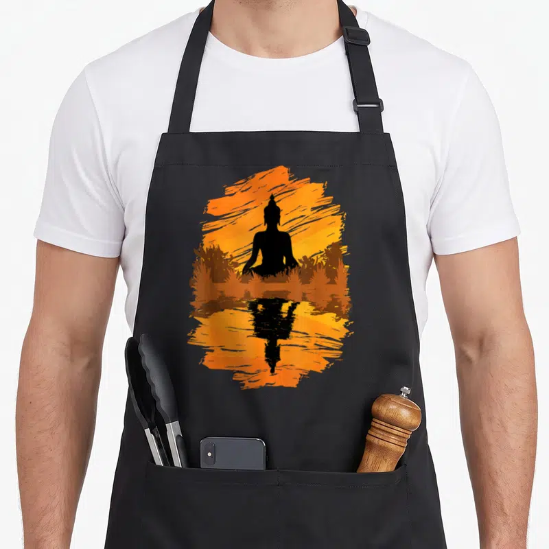 Zen asian style apron - Black Buddha silhouette meditating before orange sun with grass and water reflection — shown on a black cotton canvas apron against a white background