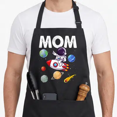 Space mother's apron - MOM text above cartoon astronaut on rocket surrounded by planets and stars — shown on a black cotton canvas apron against a white background