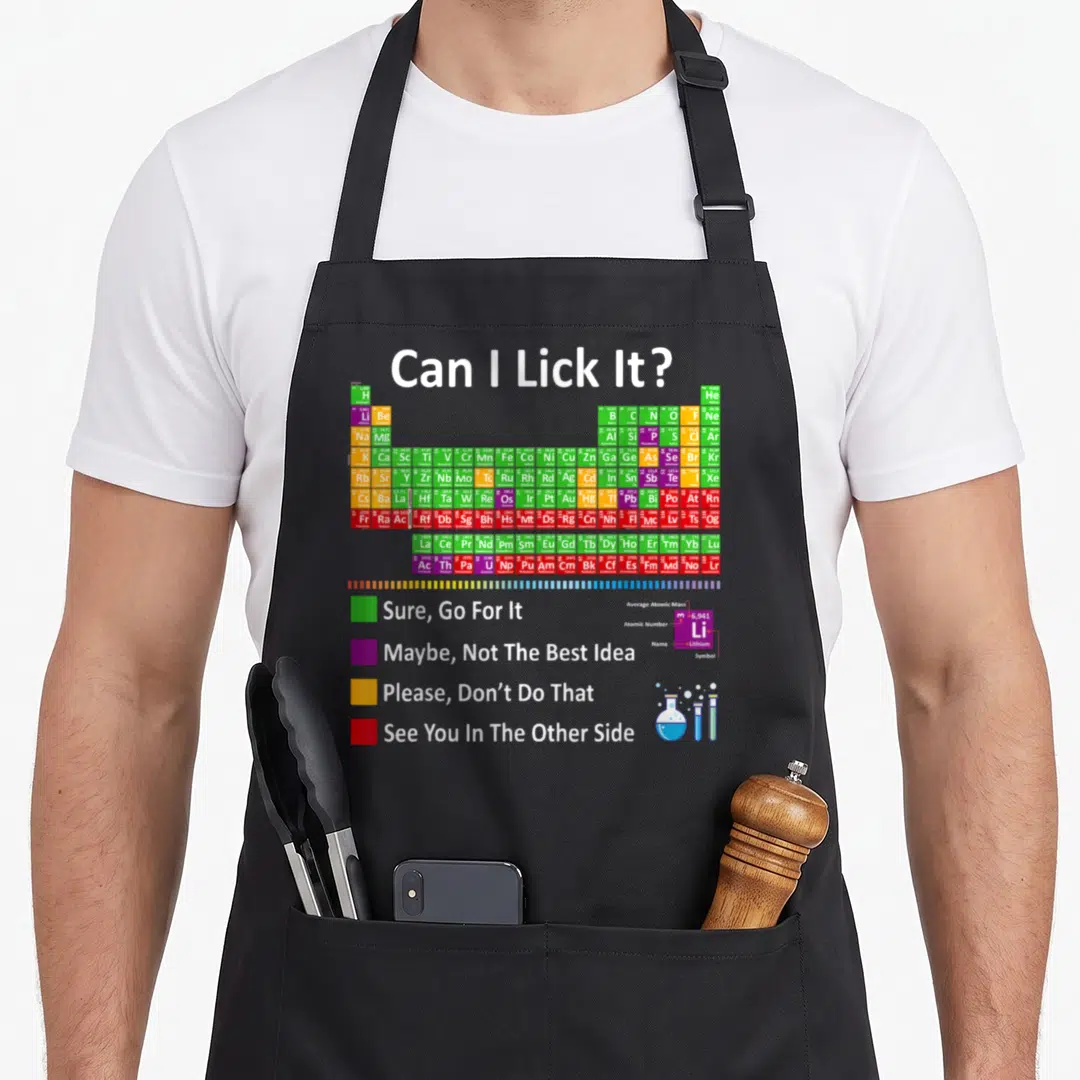 Lick chemistry apron - Periodic table with Can I Lick It text and colored safety squares next to glassware — shown on a black cotton canvas apron against a white background