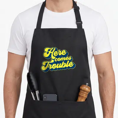 Troublemaker come and get it apron - Yellow script text Here comes Trouble outlined in blue with distressed texture — shown on a black cotton canvas apron against a white background