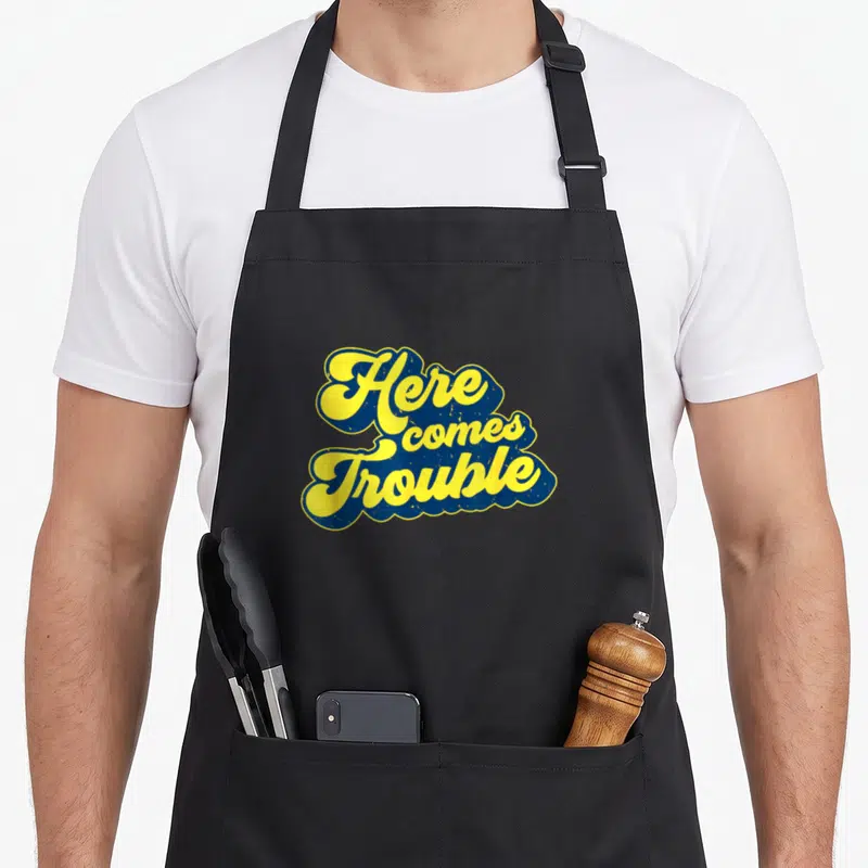 Troublemaker come and get it apron - Yellow script text Here comes Trouble outlined in blue with distressed texture — shown on a black cotton canvas apron against a white background