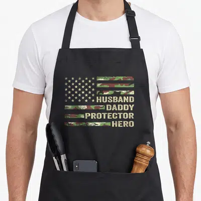 Husband camo aprons - Flag shape with stars and camo stripes reading HUSBAND DADDY PROTECTOR HERO — shown on a black cotton canvas apron against a white background