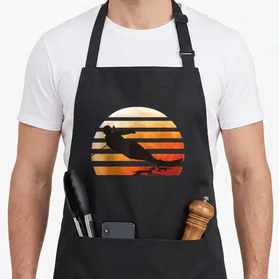 Water apron or apron - Black water skier silhouette against orange and yellow striped sunset — shown on a black cotton canvas apron against a white background