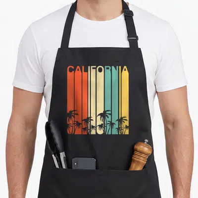 Surf 80s apron - CALIFORNIA text in red orange cream teal yellow stripes with grey palm tree silhouettes at bottom — shown on a black cotton canvas apron against a white background