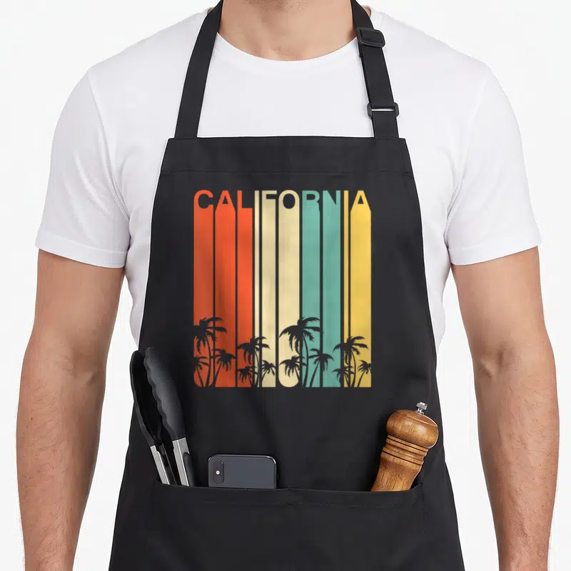 Surf 80s apron - CALIFORNIA text in red orange cream teal yellow stripes with grey palm tree silhouettes at bottom — shown on a black cotton canvas apron against a white background