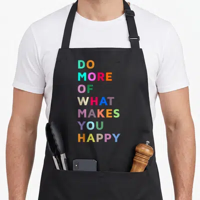 Give bahaas apron - Stacked colorful typography reading DO MORE OF WHAT MAKES YOU HAPPY in seven lines — shown on a black cotton canvas apron against a white background