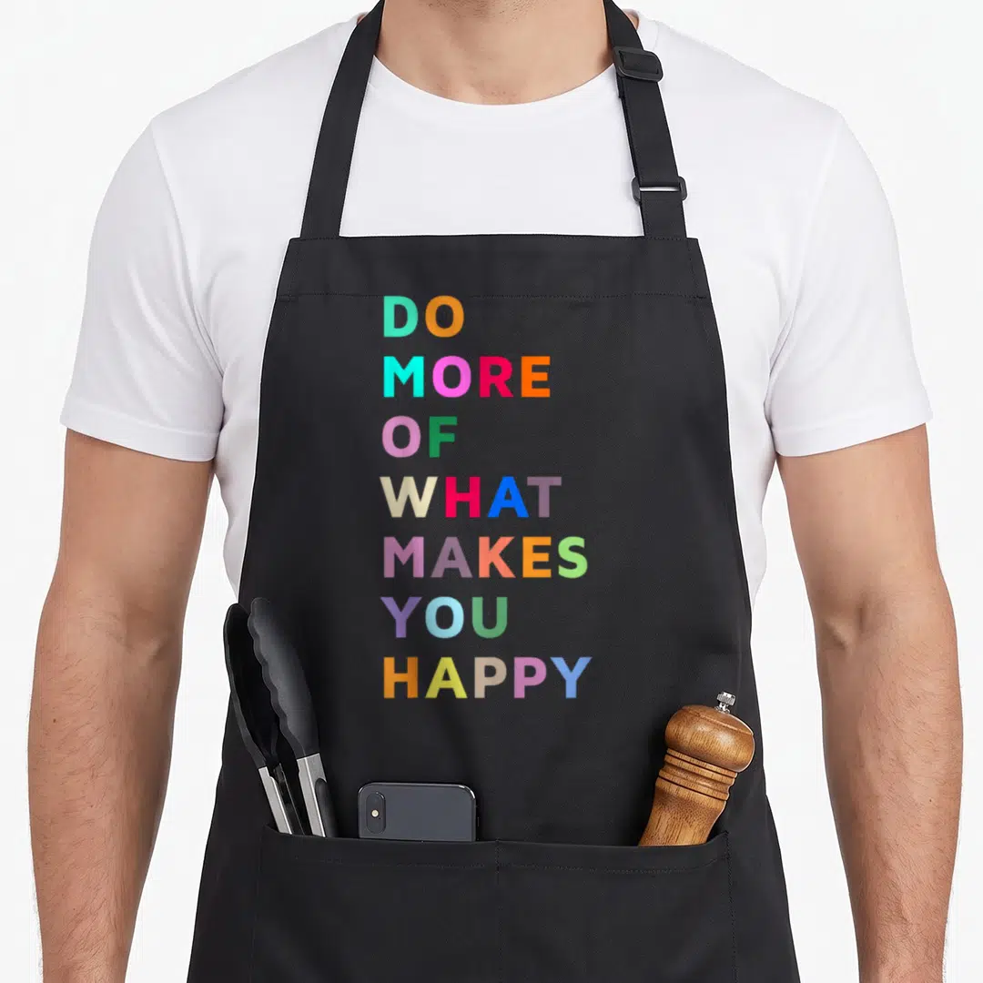 Give bahaas apron - Stacked colorful typography reading DO MORE OF WHAT MAKES YOU HAPPY in seven lines — shown on a black cotton canvas apron against a white background