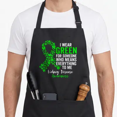 Support kidney disease awareness apron - Green shamrock awareness ribbon with white text reading I WEAR GREEN — shown on a black cotton canvas apron against a white background