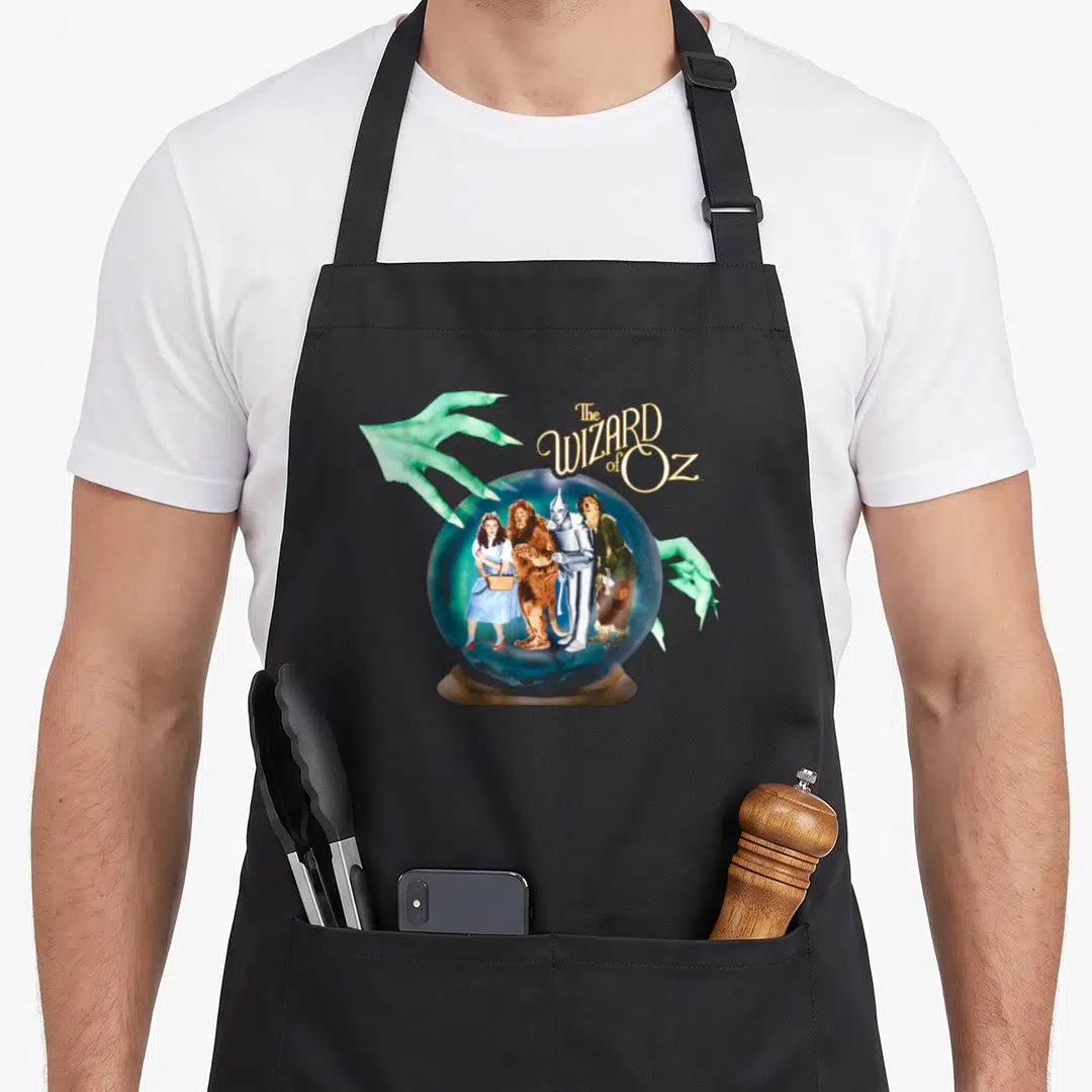 Wizard crystal apron - The WIZARD of Oz text above crystal ball with four characters and green hands — shown on a black cotton canvas apron against a white background