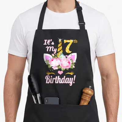 7th birthday unicorn apron - Pink unicorn face with gold 7 horn and floral center reading It's My 17th Birthday — shown on a black cotton canvas apron against a white background