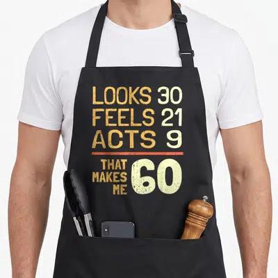 Hilarious 60th birthday apron - Orange and cream typography reading looks 30 feels 21 acts 9 that makes me 60 — shown on a black cotton canvas apron against a white background