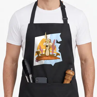Wile coyote e.a apron - Cartoon brown coyote sitting on orange rock with pink cacti and text COYOTE CARNIVOROUS — shown on a black cotton canvas apron against a white background