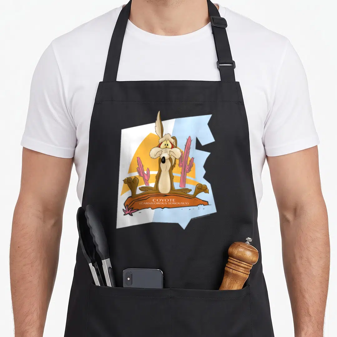 Wile coyote e.a apron - Cartoon brown coyote sitting on orange rock with pink cacti and text COYOTE CARNIVOROUS — shown on a black cotton canvas apron against a white background