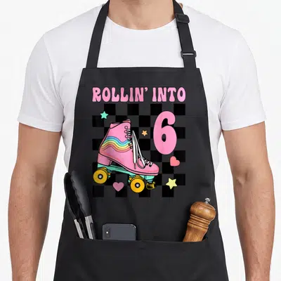 Skating 6th birthday apron - Pink roller skate and ROLLIN INTO 6 text on checkerboard background — shown on a black cotton canvas apron against a white background