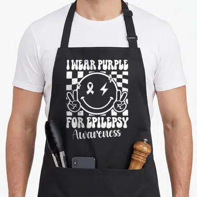 Epilepsy face apron - White smiley face with ribbon lightning bolt and peace signs on checkerboard with awareness text — shown on a black cotton canvas apron against a white background